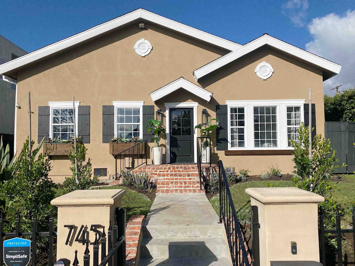 The exterior of the house features a warm beige facade with contrasting black shutters. A welcoming entrance is framed by decorative pillars and symmetrical windows. Lush greenery and well-maintained flower beds line the walkway leading to the door, set against a blue sky.