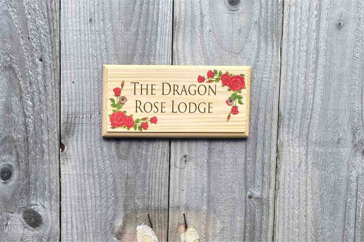 A wooden sign labeled 'The Dragon Rose Lodge' is mounted on a textured gray wooden wall, featuring decorative red roses. The sign's warm colors contrast with the natural tones of the wood, providing an inviting entrance to the lodge.