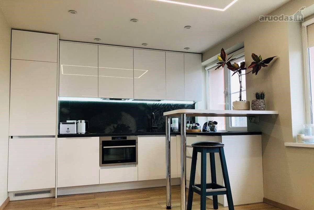 A modern kitchen is equipped with sleek white cabinetry and a dark countertop. Under-cabinet lighting illuminates the space. A small dining table with a single stool is positioned near a window, allowing natural light to enter. Appliances, including an oven, are neatly integrated into the design.