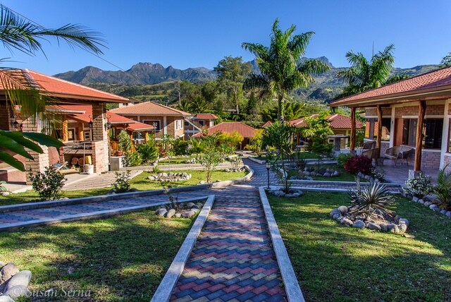 A landscaped garden area is displayed, featuring a pathway made of pavers that meanders through well-maintained grass and decorative plants. Surrounding structures with red rooftops are visible, set against a backdrop of mountains under a clear blue sky.