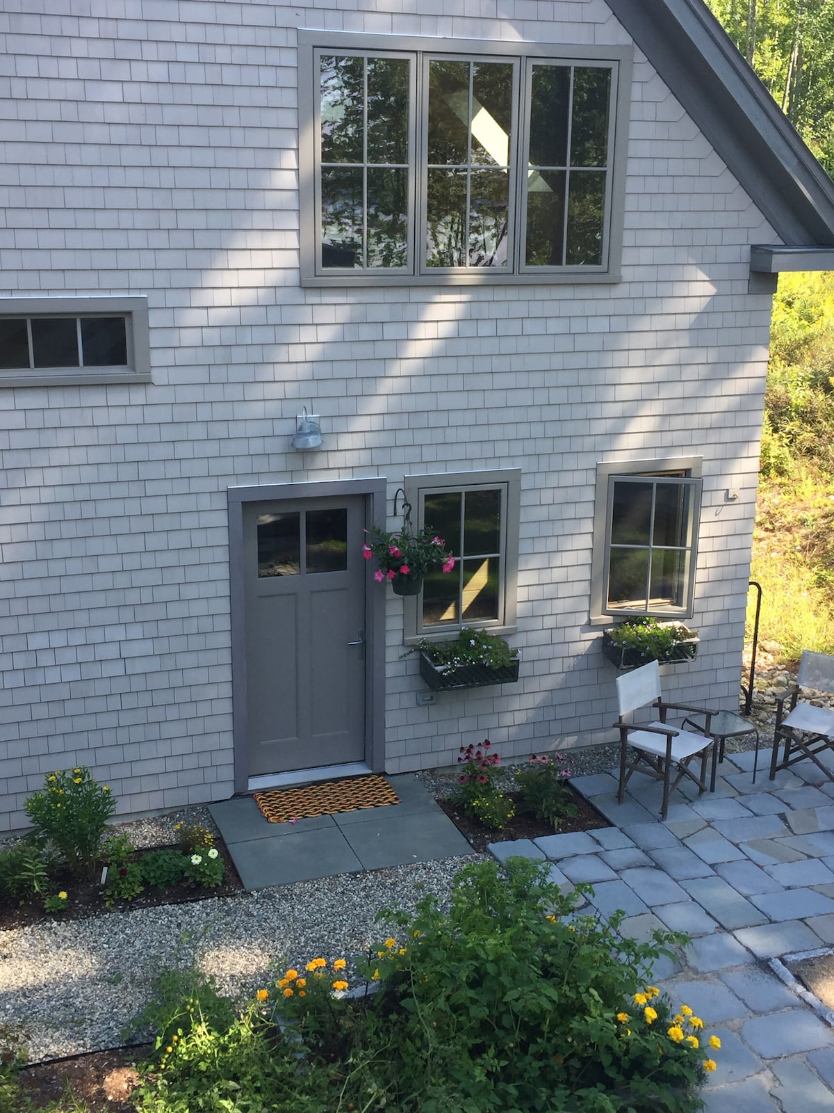 A charming entrance features a light gray door with a small decorative porch. Flower boxes filled with colorful plants adorn the windows. A slate pathway leads to the door, surrounded by a garden with vibrant flowers and greenery. Two chairs are positioned nearby for outdoor seating.