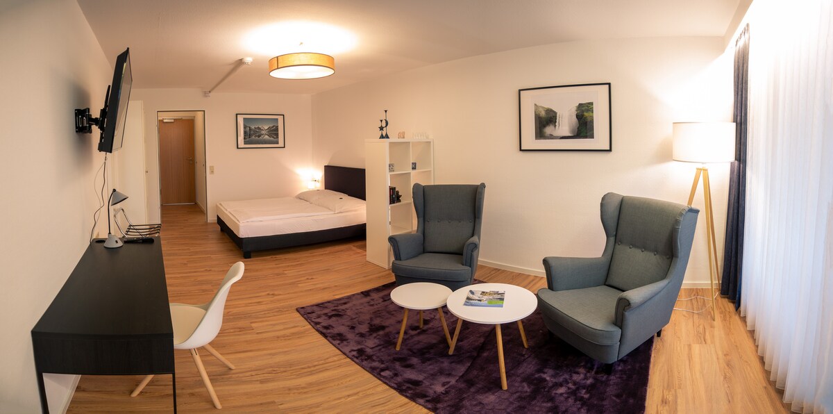 A spacious room features a comfortable bed and a seating area with two armchairs around a small coffee table. A desk is positioned against the wall, and a television is mounted above it. Natural light enters through large windows covered with sheer curtains.