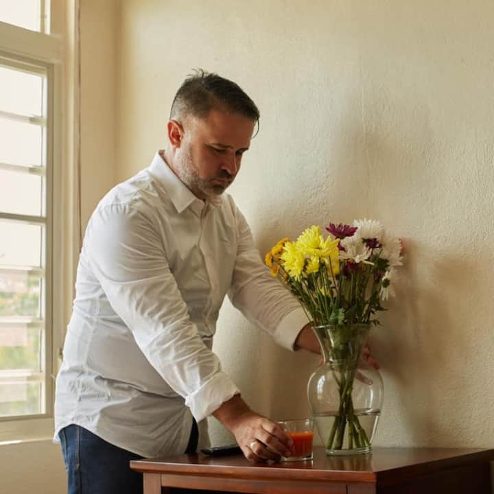 A person with a gray beard wearing a white button-down shirt places a vase of flowers on a table.