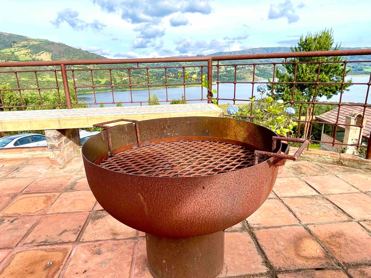 An outdoor barbecue grill sits on a tiled patio, overlooking a serene body of water. Views of lush hills are visible in the background, adding to the scenic ambiance. The grill features a circular design with a mesh top for cooking.