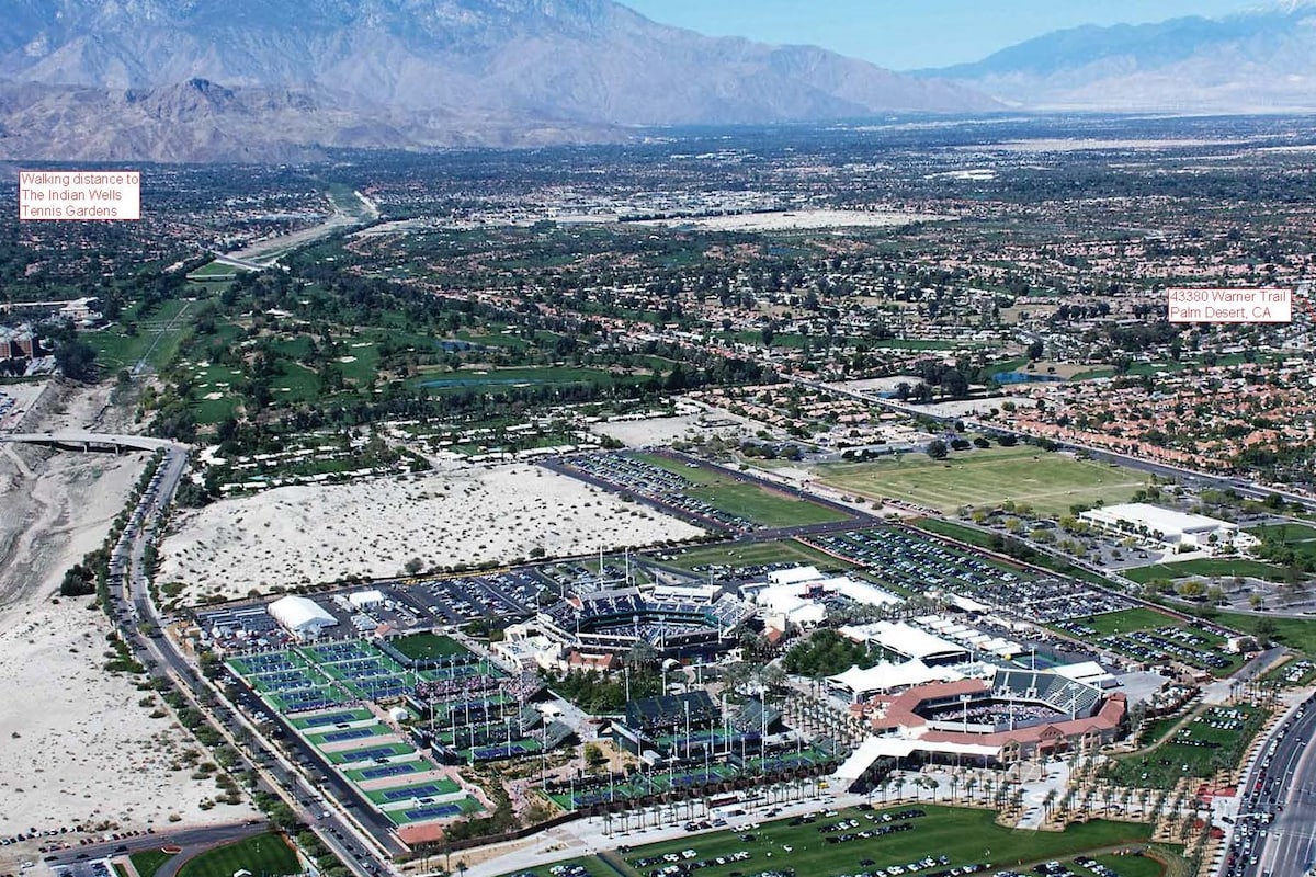 An aerial view captures the surrounding area of the Indian Wells Tennis Gardens, showcasing expansive parking lots, lush greenery, and the nearby mountain range. The vast desert landscape is visible, illustrating the proximity to the Coachella Valley's natural beauty.