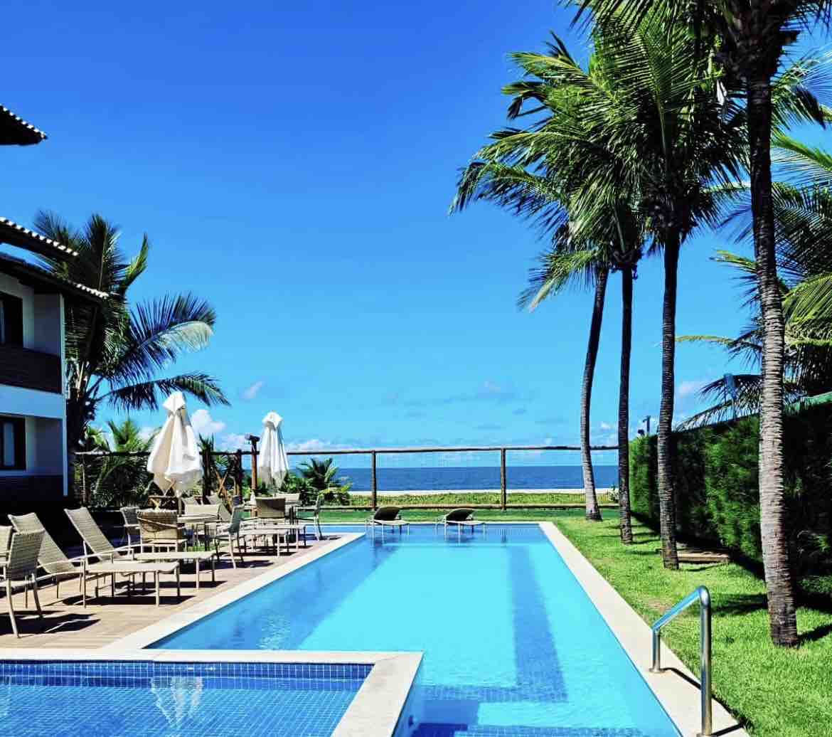 A pool area features a clear blue swimming pool surrounded by lounge chairs. Palm trees provide shade, and the ocean is visible in the background under a bright blue sky.