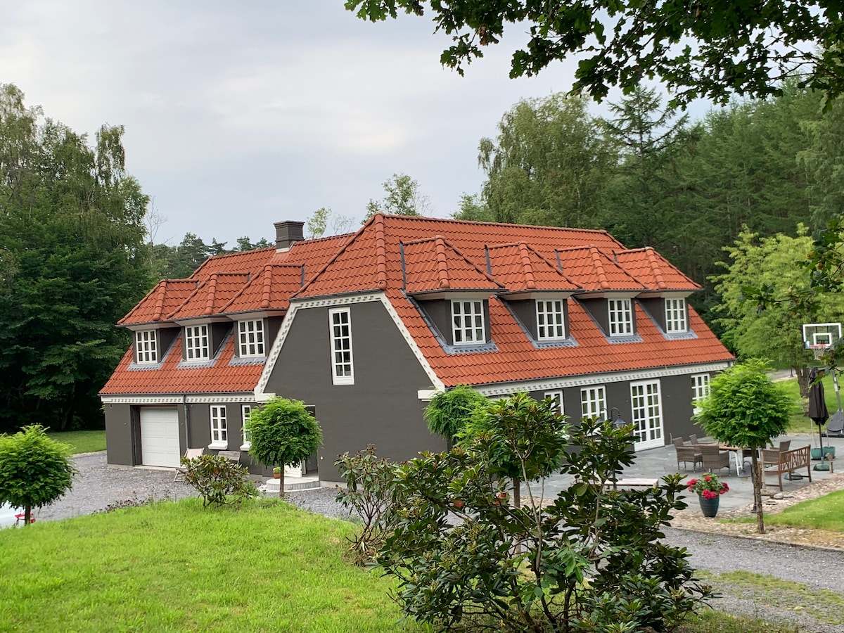 The retreat features a charming two-story structure with a unique mix of dark gray and red accents. A sloped, tiled roof complements the windows that allow natural light. The surrounding greenery includes neatly trimmed bushes and open spaces, enhancing the peaceful outdoor environment.