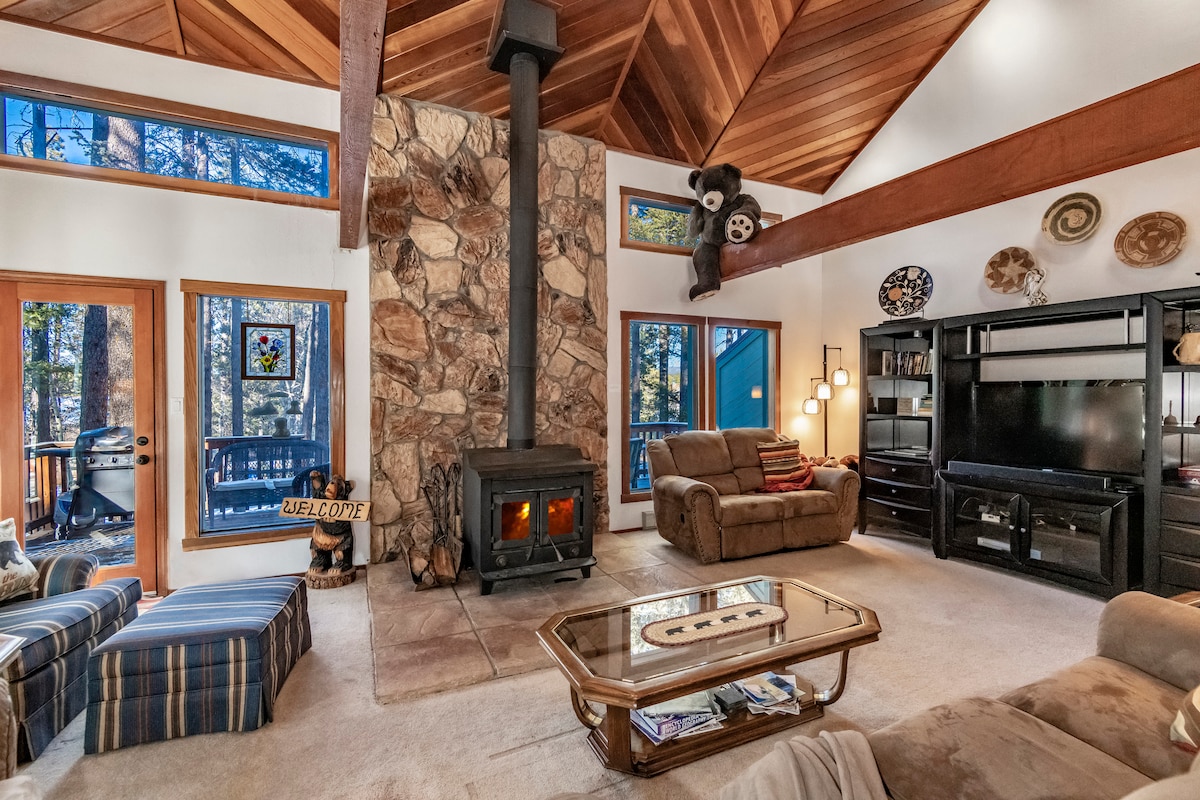 A spacious living area features a large stone fireplace as the focal point, surrounded by comfortable seating options. Large windows provide views of the woods, while natural light fills the space, complemented by wooden beams on the ceiling.