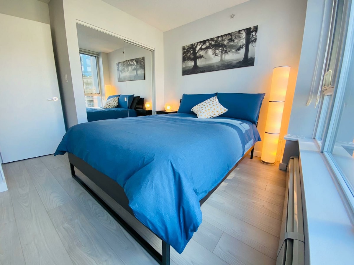 The bedroom features a spacious bed covered in a blue duvet, complemented by a decorative pillow. Soft lighting is provided by lamps on either side of the bed, and an accent mirror reflects the natural light from the window, enhancing the room's bright atmosphere.