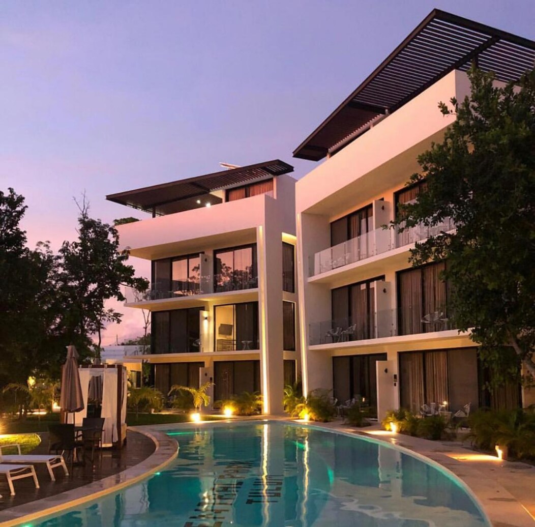 Two modern buildings are positioned alongside a curved pool, with greenery surrounding the area. Soft lighting enhances the exterior, highlighting large glass windows and balconies. A lounge area is visible, featuring umbrellas and patio furniture against a tranquil evening sky.