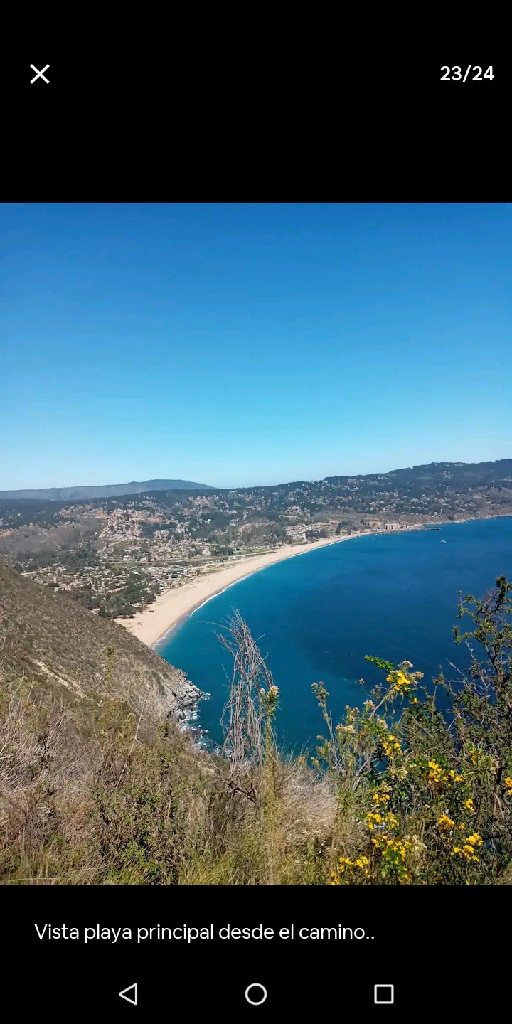 A panoramic view of the main beach is captured from an elevated path. The coastline curves gently, with golden sands meeting the blue waters of the ocean. Rolling hills and greens surround the beach, while a clear sky provides a serene backdrop.