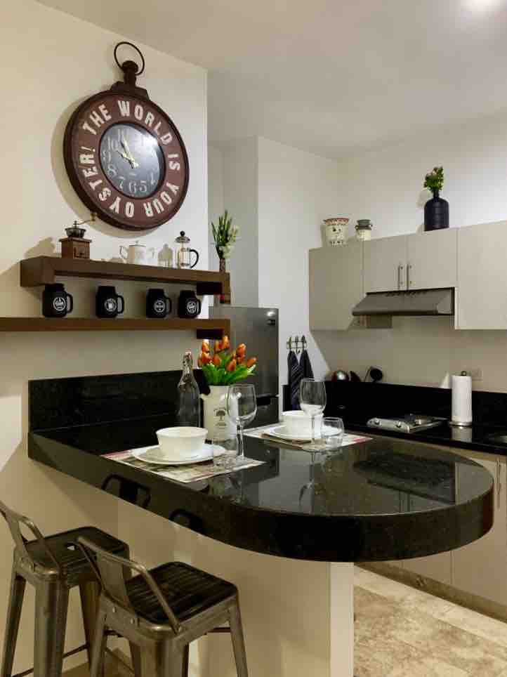 A modern kitchen features sleek cabinetry and a black countertop, accented by a wall clock and decorative plants. A dining area is depicted with two place settings and glasses, inviting a comfortable dining experience.