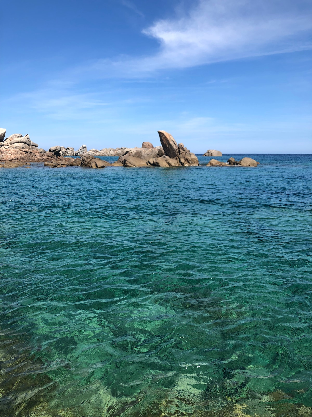 The image captures a clear blue sea, revealing rocky formations above and below the water's surface. Sunlight reflects off the calm water, creating a shimmering effect. The horizon showcases a clear sky with soft clouds, enhancing the serene coastal atmosphere.
