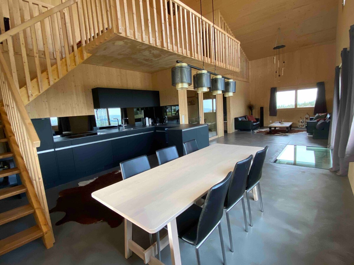 An open living area is showcased, featuring a large dining table with black chairs. A modern kitchen with a sleek black finish occupies one side. Large windows allow natural light to fill the space, with a cozy seating area visible in the background. Stairs lead to a lofted area above.