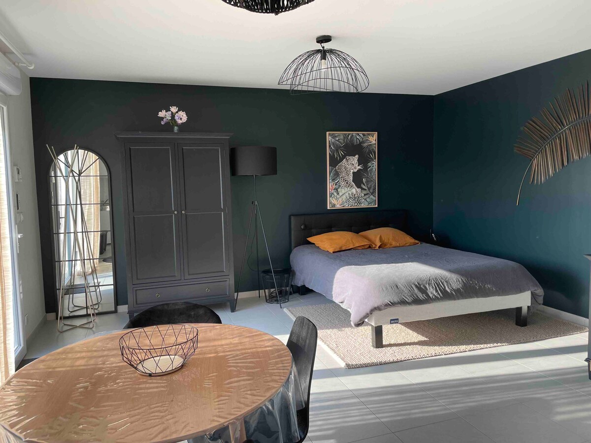 An open living space features a double bed with neutral bedding, accompanied by decorative pillows. A round dining table with a metallic finish is positioned nearby, alongside an elegant wardrobe. A floor lamp and wall art contribute to the modern decor, while ample natural light enters through large windows.