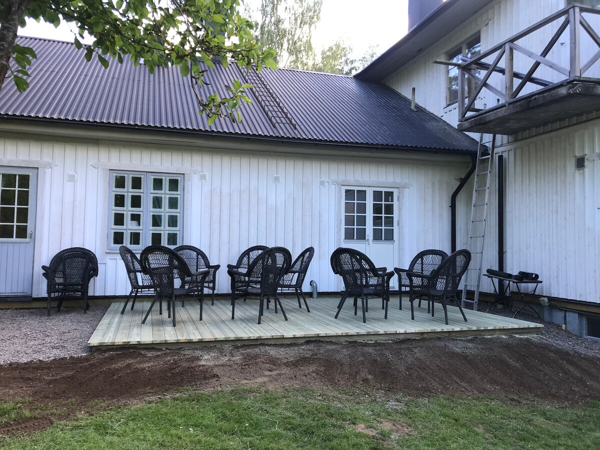 An outdoor deck area is shown, featuring several black wicker chairs arranged for seating. The deck is situated next to a white wooden building, with large windows allowing for natural light. Grass surrounds the deck, providing a spacious outdoor environment.