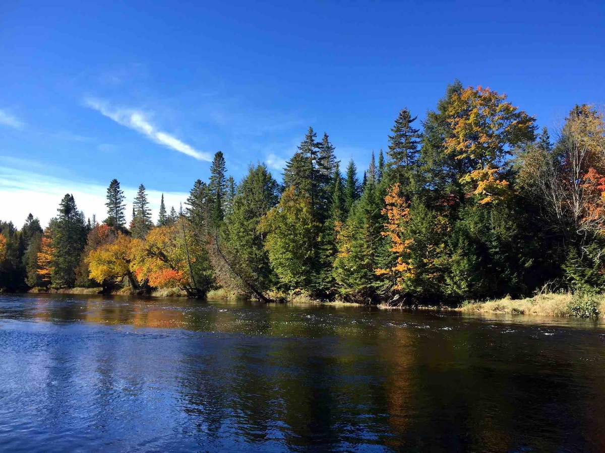 The tranquil river is bordered by vibrant trees displaying autumn colors, ranging from deep oranges to bright yellows. A clear blue sky reflects softly on the water's surface, enhancing the natural beauty of the surrounding landscape.