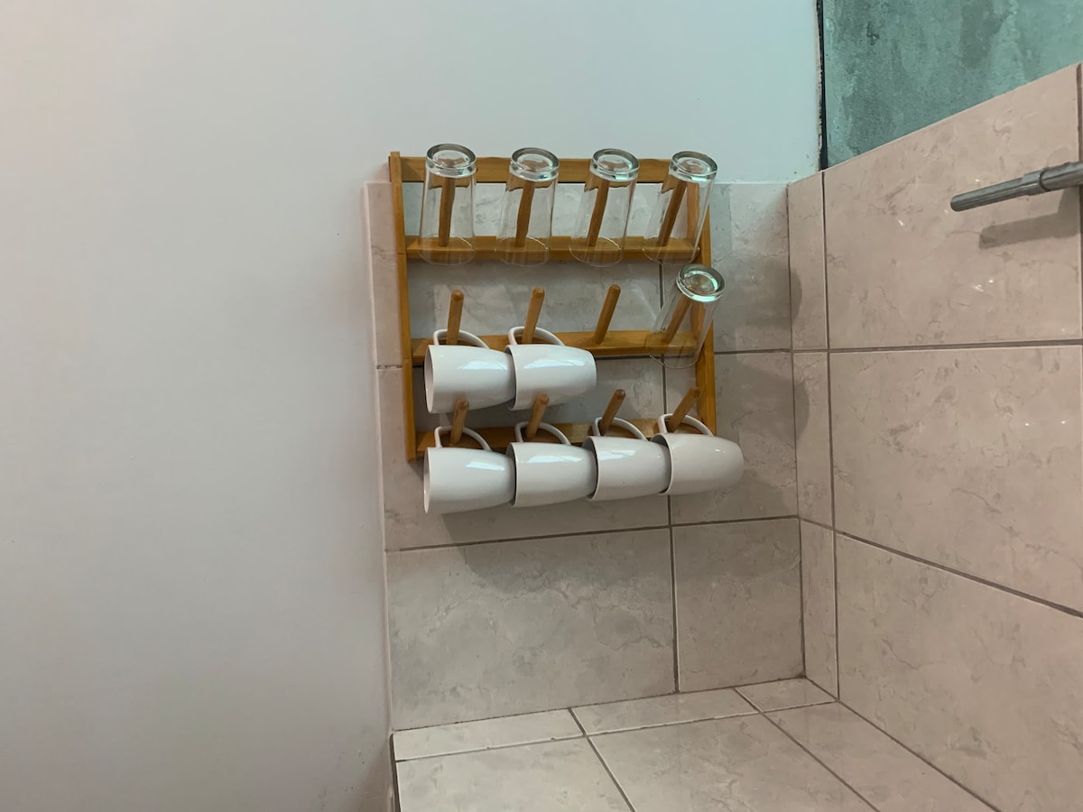 A wooden rack is mounted on the wall, displaying a total of eight white cups and three glasses. The cups are neatly arranged, showcasing a simple design, while the tiles below add a neutral backdrop.