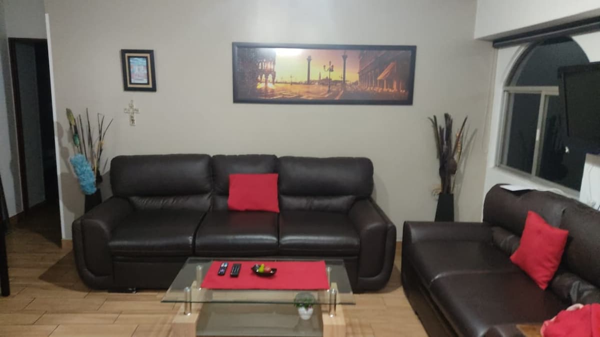 A modern living area features two dark leather sofas adorned with red cushions. A glass coffee table sits in the center, displaying a decorative tray and remote controls. A large artwork is mounted on the wall, complemented by decorative vases placed in the corners.