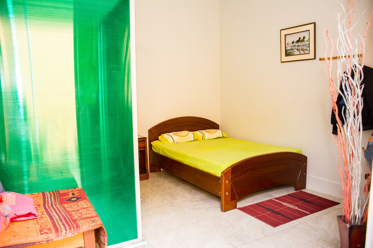 A sleeping area is presented with a wooden bed featuring a vibrant green blanket. Soft lighting enhances the simplicity of the space. A small table is positioned nearby, accompanied by a decorative plant in a simple vase.