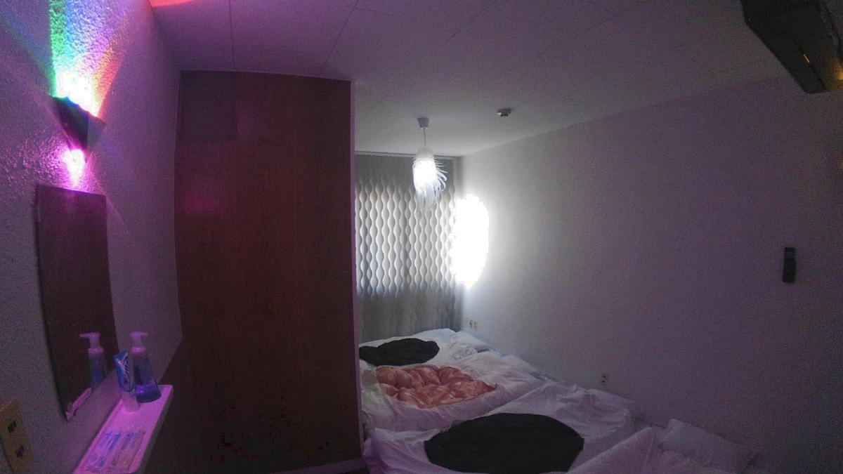 A spacious room features two beds with layered bedding, positioned against a smooth wall. Soft lighting from a geometric fixture creates an ambient glow, while a window covered with patterned shades allows gentle daylight to filter through, enhancing the minimalistic decor.