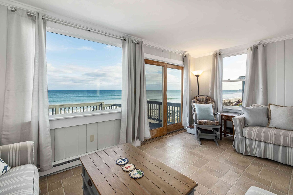 A light-filled living area features large windows offering panoramic ocean views. Soft gray curtains frame the windows, while a wooden coffee table sits at the center. Comfortable seating arrangements include an armchair and a side table, creating a relaxing space overlooking the beach.
