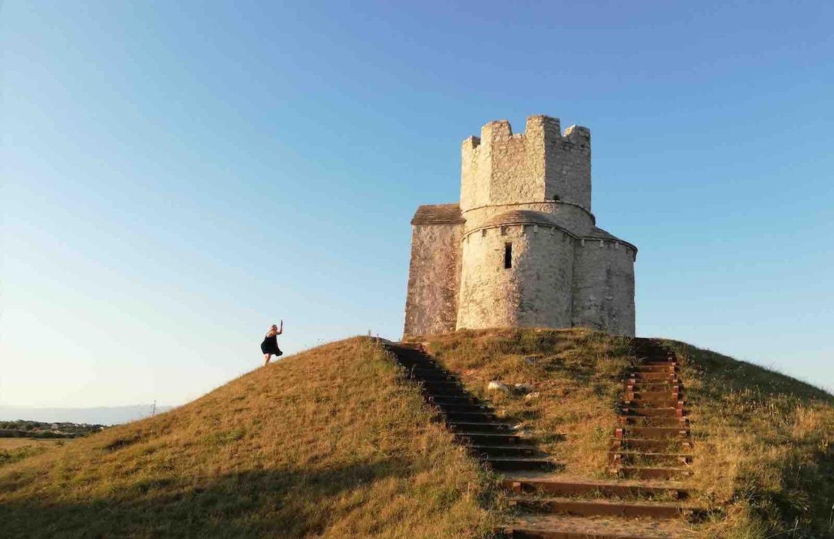 A historical stone tower is positioned atop a grassy hill, surrounded by a clear blue sky. A pathway with a few stone steps leads up to the structure, while a figure is seen in the foreground, interacting with the landscape.