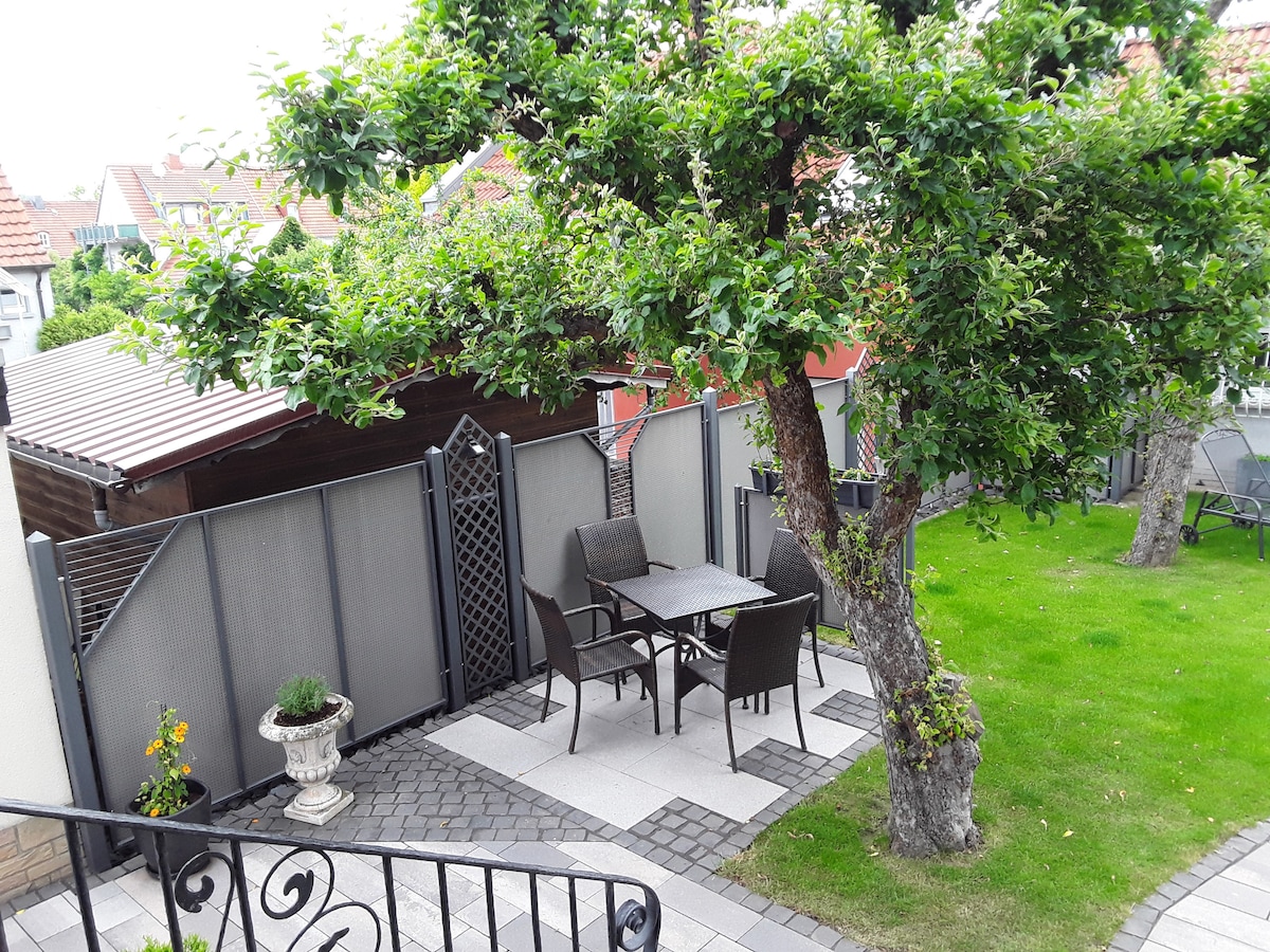 An outdoor seating area is featured, surrounded by greenery. A square table with four chairs is positioned on a stone-paved surface. A small potted plant adds a touch of nature, while a large tree provides shade nearby. A fence encloses the area for privacy.