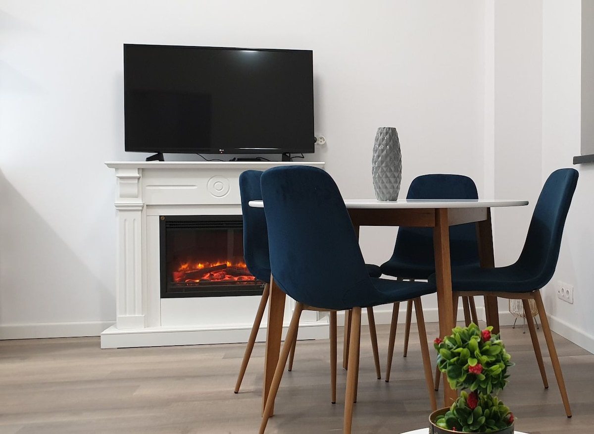 A modern dining area features a round white table surrounded by four blue chairs. A sleek television is mounted above a white fireplace with an electric flame, adding a cozy touch. A small potted plant sits on the table, contributing to the overall ambiance.