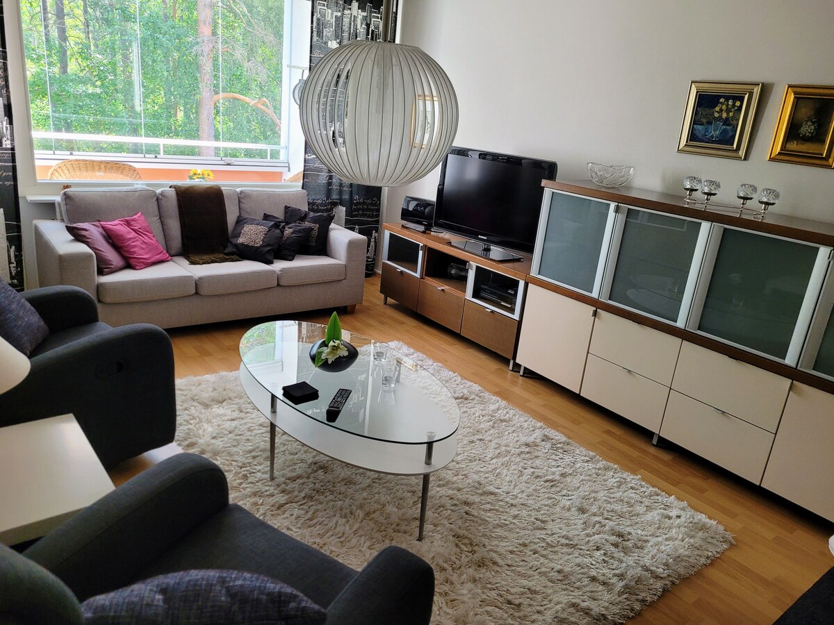 A cozy living room area features a light-colored sofa arranged with decorative pillows, complemented by two dark armchairs. A round table sits in the center, accompanied by a soft area rug. Natural light is welcomed through a large window showcasing greenery outside.