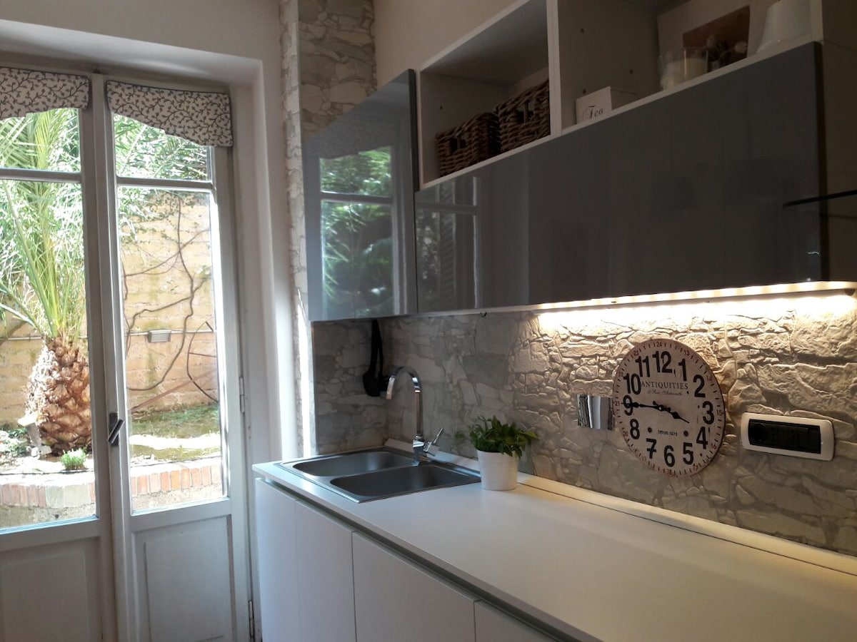 A modern kitchen is presented with sleek, gloss-finished cabinetry and a spacious countertop. A wall clock adds a functional element to the decor. Natural light floods in through a glass door leading to a private garden, enhancing the fresh ambiance of the space.