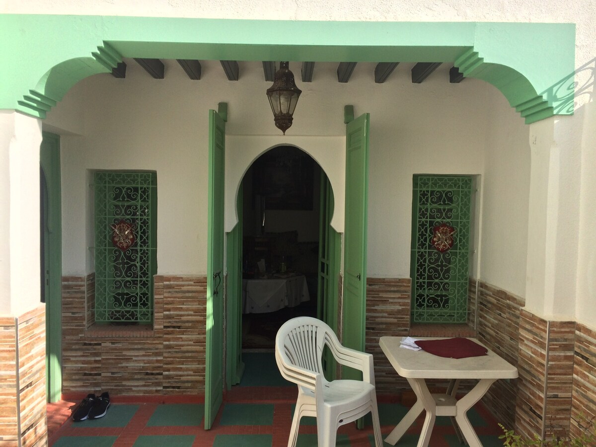 An outdoor seating area features a simple white table with a maroon placemat and a white chair. Two green doors open into the space, framed by decorative arches. Intricate designs are seen in the adjacent windows, with tiled accents visible on the walls.