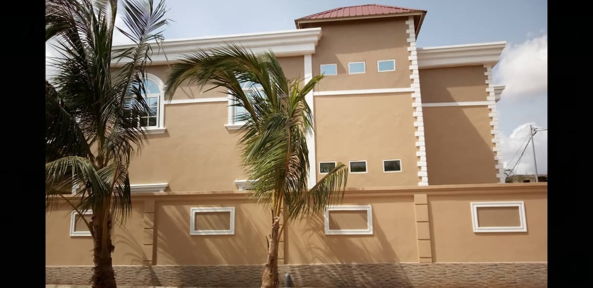 The exterior of a three-bedroom apartment is highlighted, showcasing a modern design with light brown walls and white decorative trim. Several windows are visible, providing natural light. Palm trees are positioned in the foreground, adding greenery to the setting.