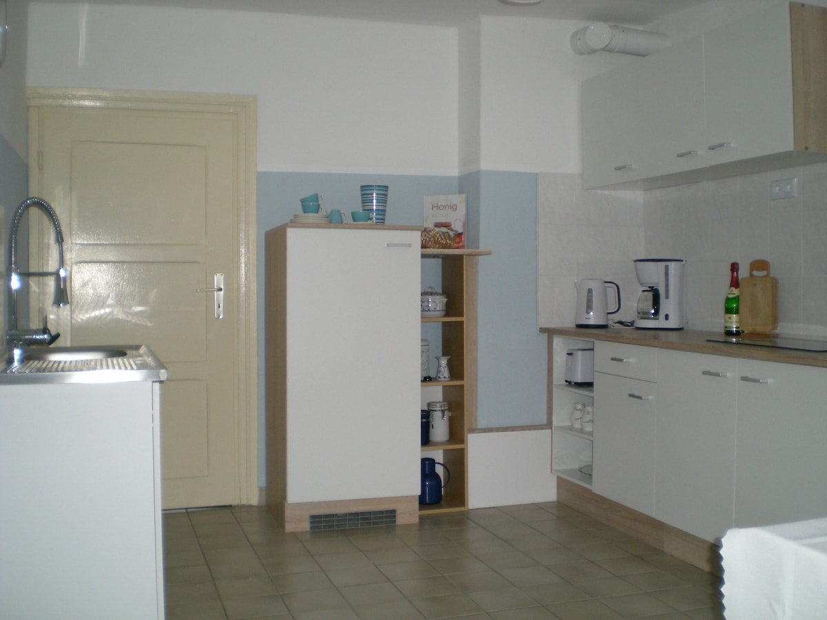 The kitchen area features modern cabinetry with a clean, functional layout. A refrigerator and sink are positioned alongside shelves containing dishware and appliances. Countertop appliances, including a coffee maker and kettle, are visible. Natural light brightens the space through a nearby door.