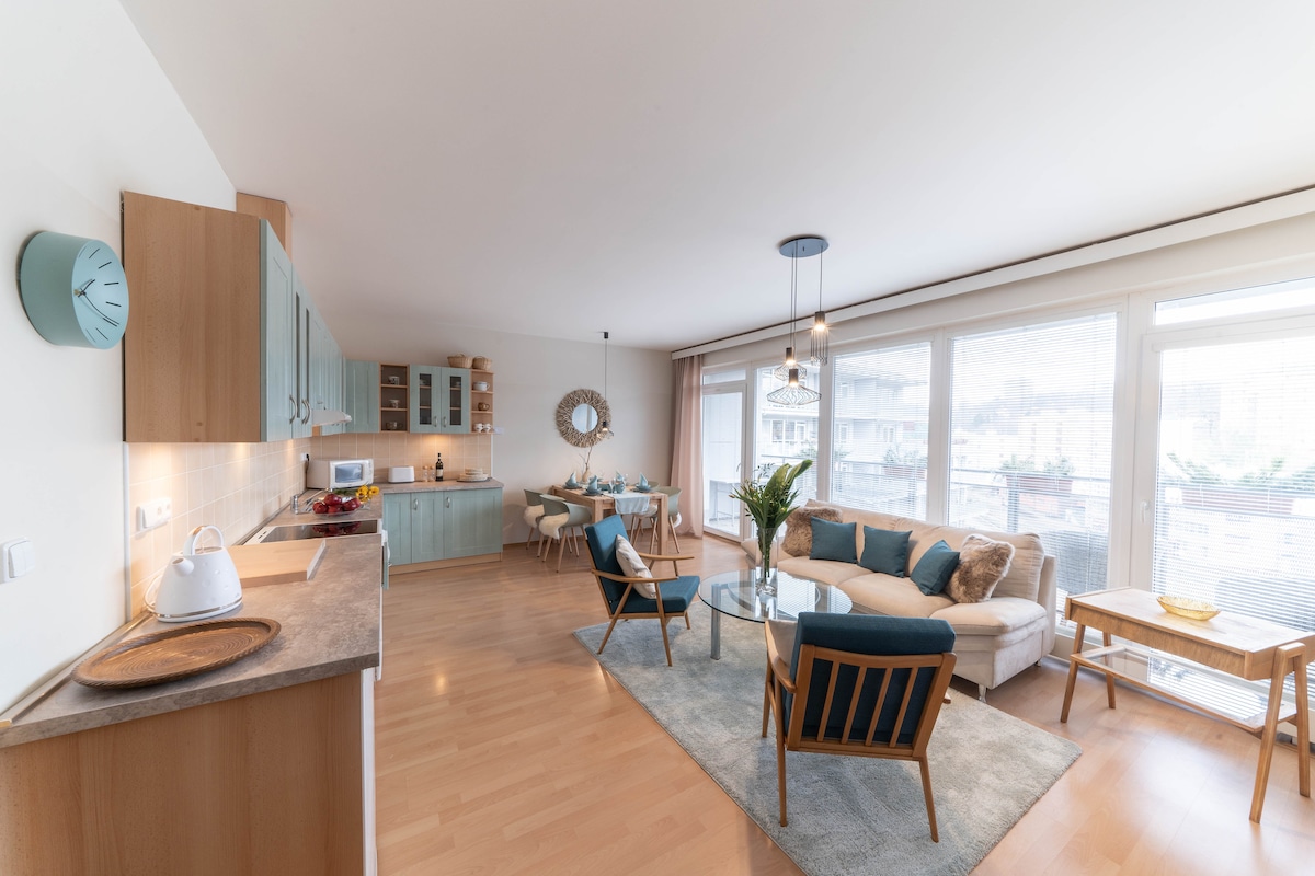 A modern open-plan living area is showcased, featuring a comfortable seating arrangement with a light-colored sofa and stylish chairs. A kitchen area with wooden cabinetry and a dining table for four is visible. Natural light fills the space, enhanced by large windows.