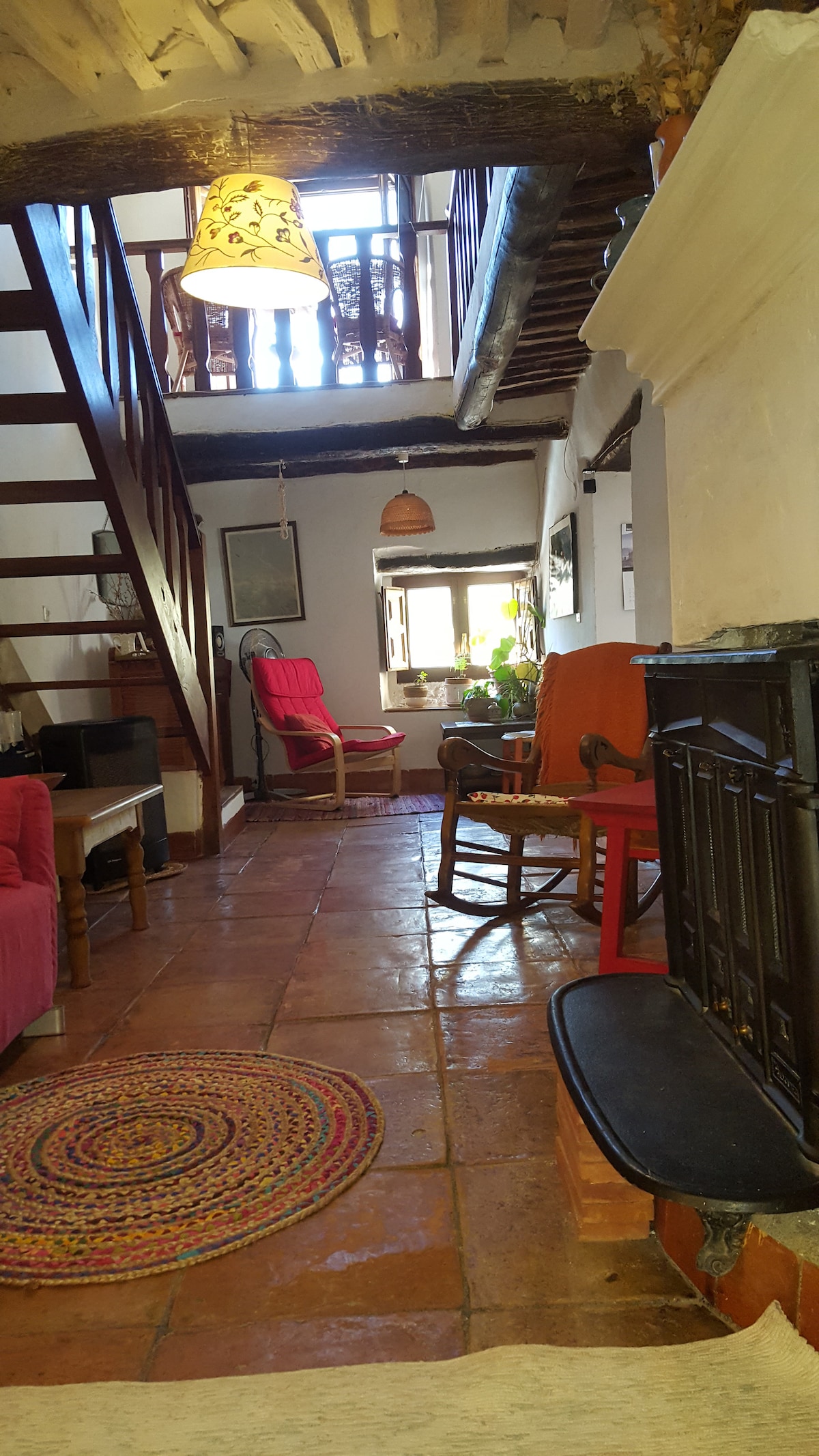 A welcoming interior features terracotta tiled flooring, complemented by colorful furnishings. A bright red chair and a rocking chair are positioned near a window with plants. Wooden beams add character, while a circular rug offers a soft touch to the living space.