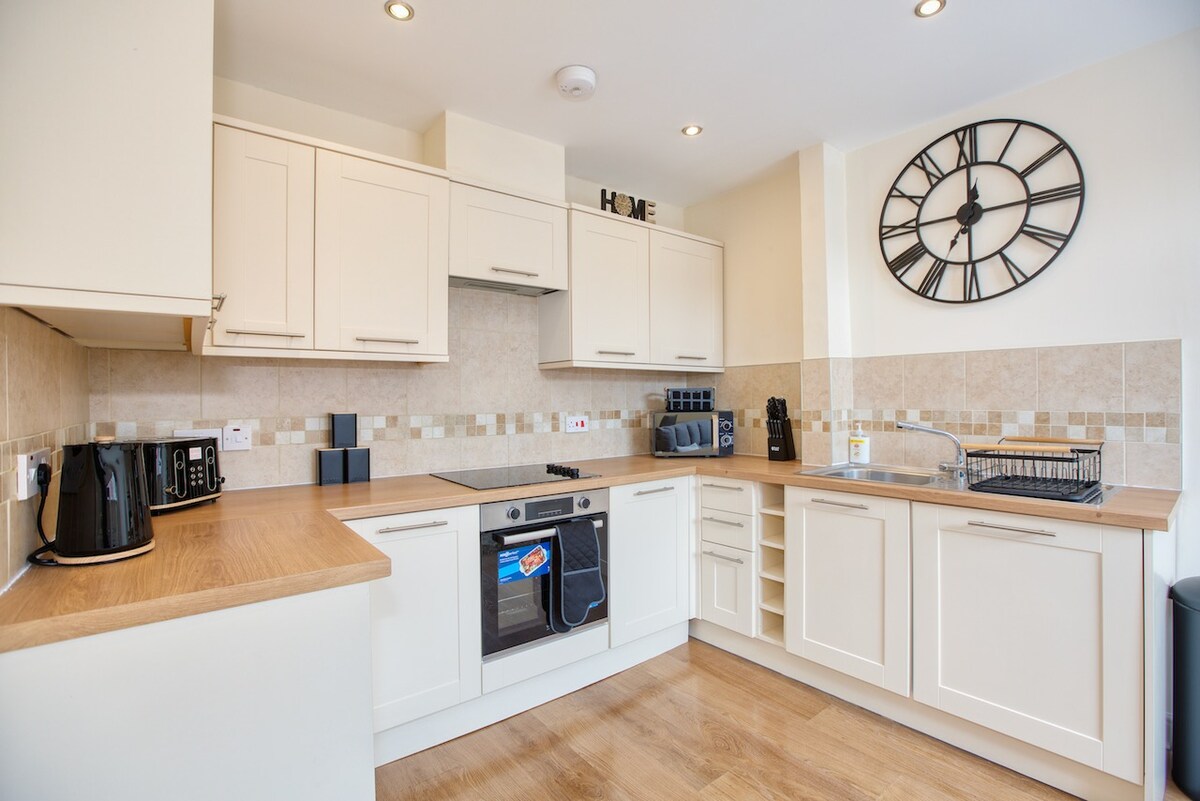 A well-equipped kitchen is presented, featuring modern appliances including an oven and microwave, sleek cabinetry, and a spacious wooden countertop. A large wall clock adds a functional touch, while ample storage space is available for kitchen essentials.