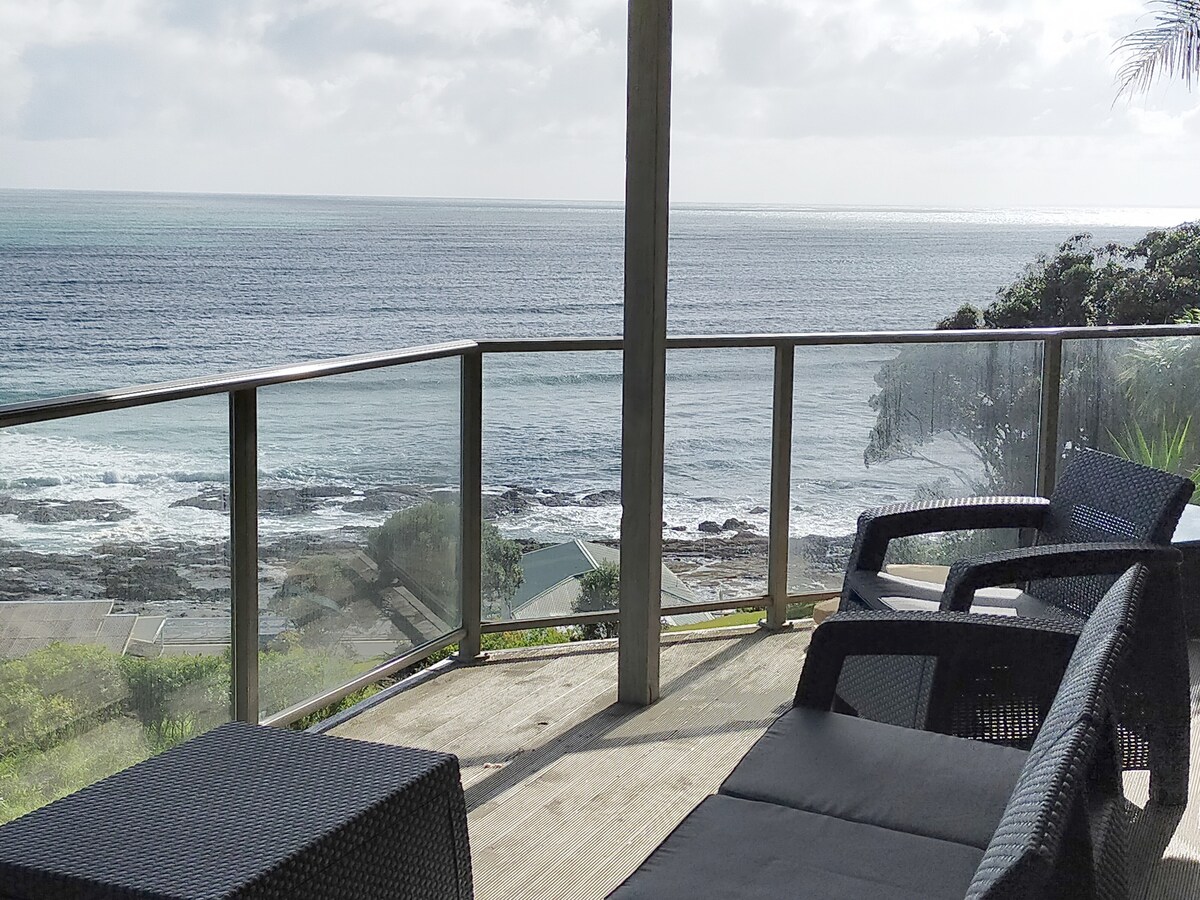 A serene balcony is presented, featuring comfortable seating arranged to face the ocean. The glass railing offers unobstructed views of the water and shoreline. Natural light filters through, enhancing the space where the sound of waves can be heard in the background.