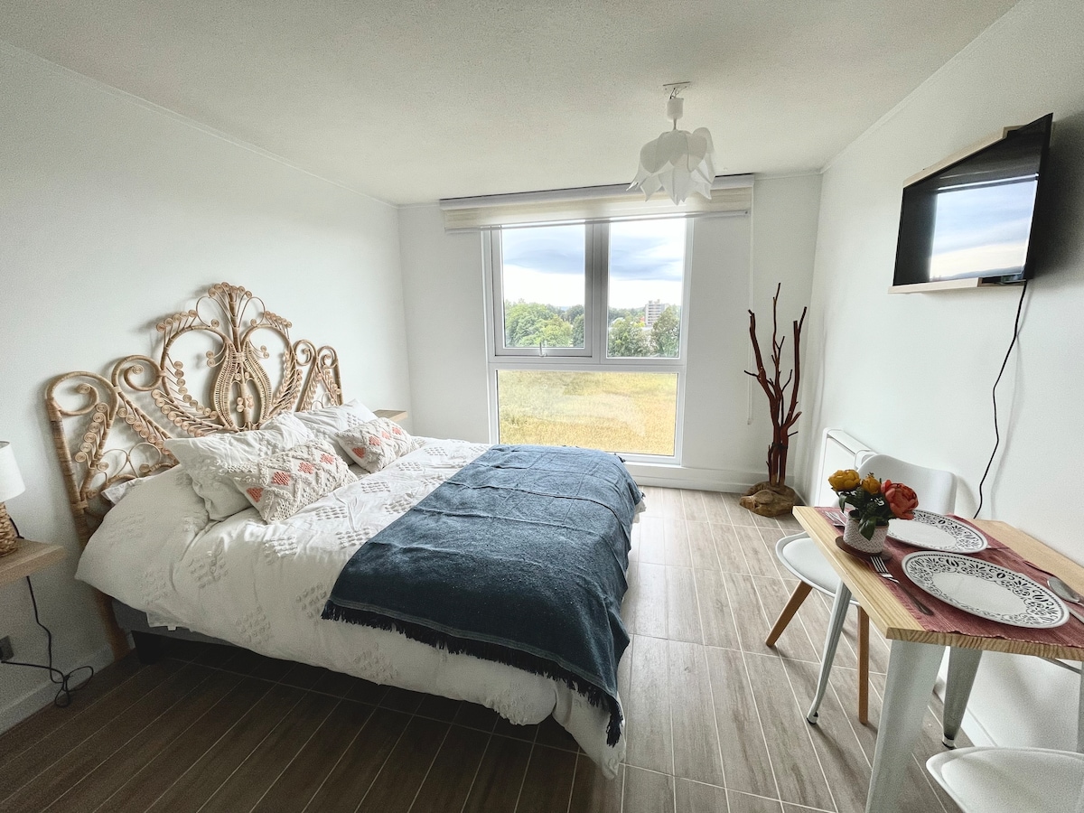 A bright bedroom features a spacious bed adorned with textured linens and decorative pillows. Natural light streams through large windows, offering views of the surrounding greenery. A small dining table sets nearby, complemented by a wall-mounted television and elegant décor details, enhancing the modern aesthetic.