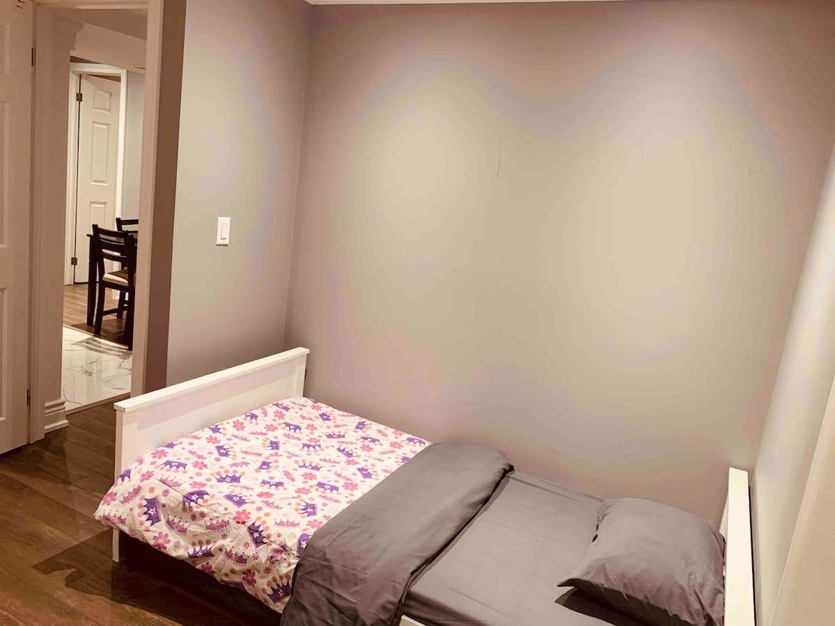 A simple sleeping arrangement is presented with a white frame bed featuring a floral-patterned blanket and a dark gray sheet. The surrounding walls are painted in a muted gray tone, while warm wood flooring complements the space. A doorway and dining chairs are visible in the background.