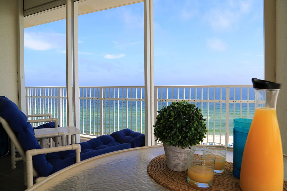 A coastal balcony is captured, featuring a glass table set with orange juice and decorative candles. Comfortable seating provides a view of the ocean, where blue waters meet a clear sky, framed by the balcony's expansive glass railing.