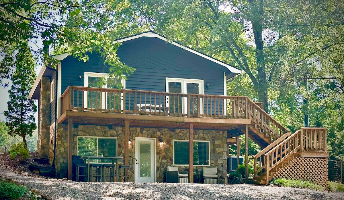 The exterior of the cottage is shown, featuring a two-story structure with a mix of wooden and stone elements. A large wraparound deck is present, surrounded by trees, providing a serene woodland setting. Multiple patio doors lead to the upper deck area, ideal for outdoor relaxation.