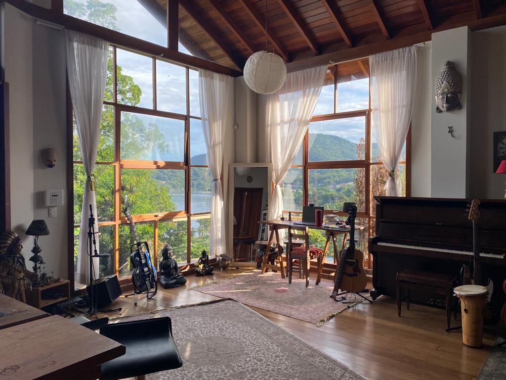 An inviting living space is adorned with large windows that offer panoramic views of the surrounding nature and lake. Wooden beams highlight the ceiling, while a piano and musical instruments are arranged thoughtfully in the room. Soft light filters through sheer curtains, enhancing the serene environment.