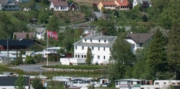 A white, two-story building is framed by greenery and residential homes. A flag of Norway is displayed prominently in front, alongside a well-maintained lawn. Various vehicles are parked nearby, contributing to the community setting.