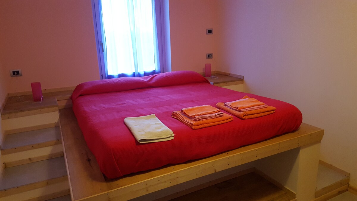 A spacious bed with a bright red coverlet is positioned on a raised platform. Neatly arranged towels in shades of yellow and orange are placed at the foot of the bed. Natural light enters through a window, adding warmth to the peach-colored walls.