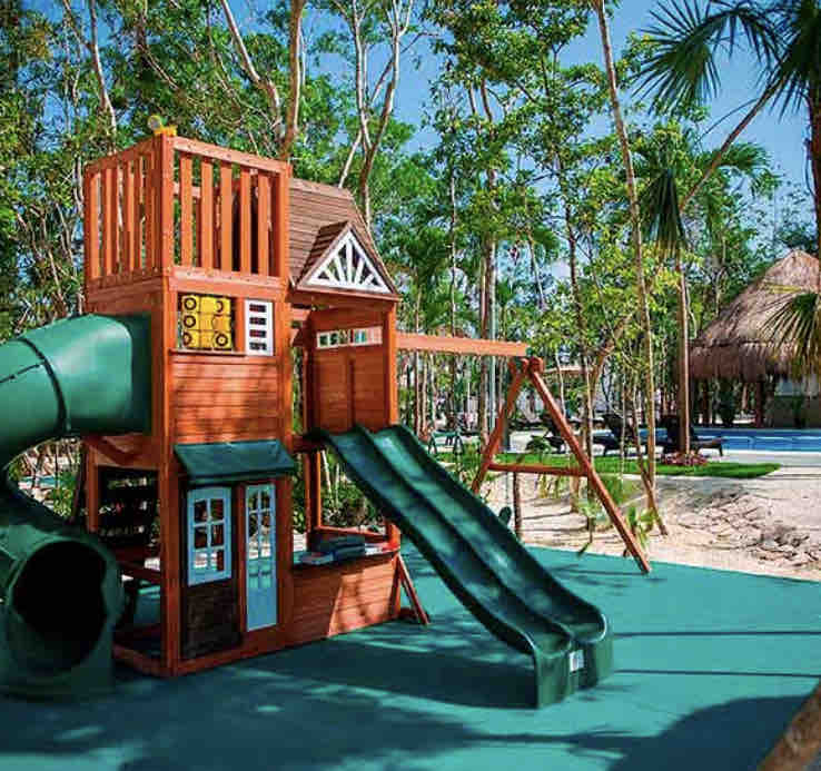 A children’s playground is set within a natural landscape, featuring a wooden structure with a slide and a climbing area. Nearby, swings are visible, and vibrant green trees surround the play area, creating an inviting space for outdoor activities.
