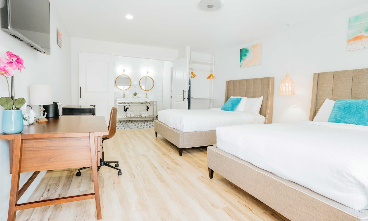 A modern guest room features two spacious beds with neutral-toned headboards. A wooden desk and chair sit beside a large mirror, illuminated by two circular light fixtures. A bright decor element is added by wall art and a potted plant. Light wood flooring completes the design.