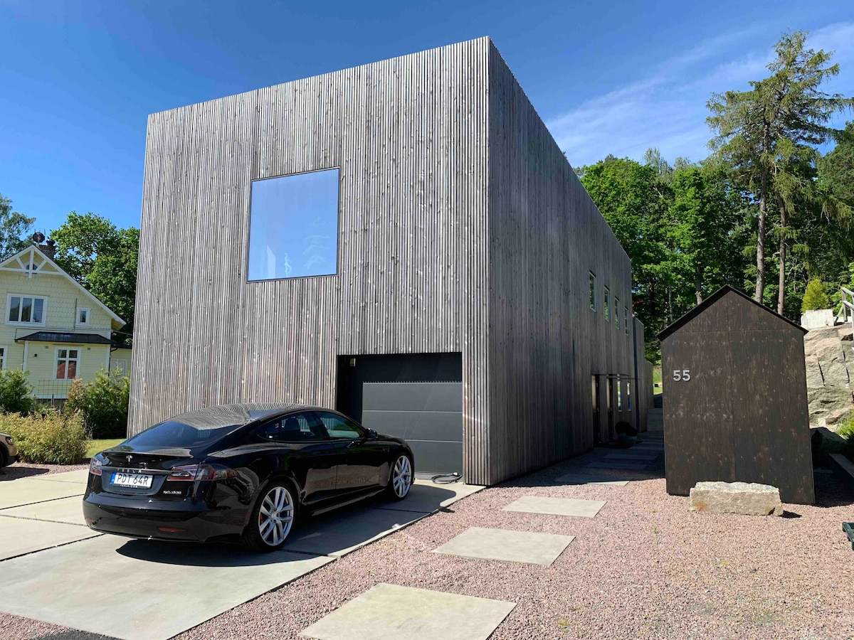 A modern villa with a wooden exterior is surrounded by greenery. Large windows are present on the façade, allowing natural light to illuminate the interior. A black car is parked in front of a garage, with a paved driveway leading to the entrance.