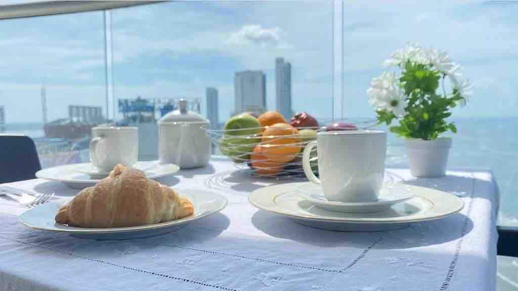 A breakfast setting is displayed on a table with a scenic view of the ocean. A croissant rests on a white plate, accompanied by a cup and saucer. A fruit bowl filled with various fruits and a small flower arrangement are positioned nearby, enhancing the inviting atmosphere.