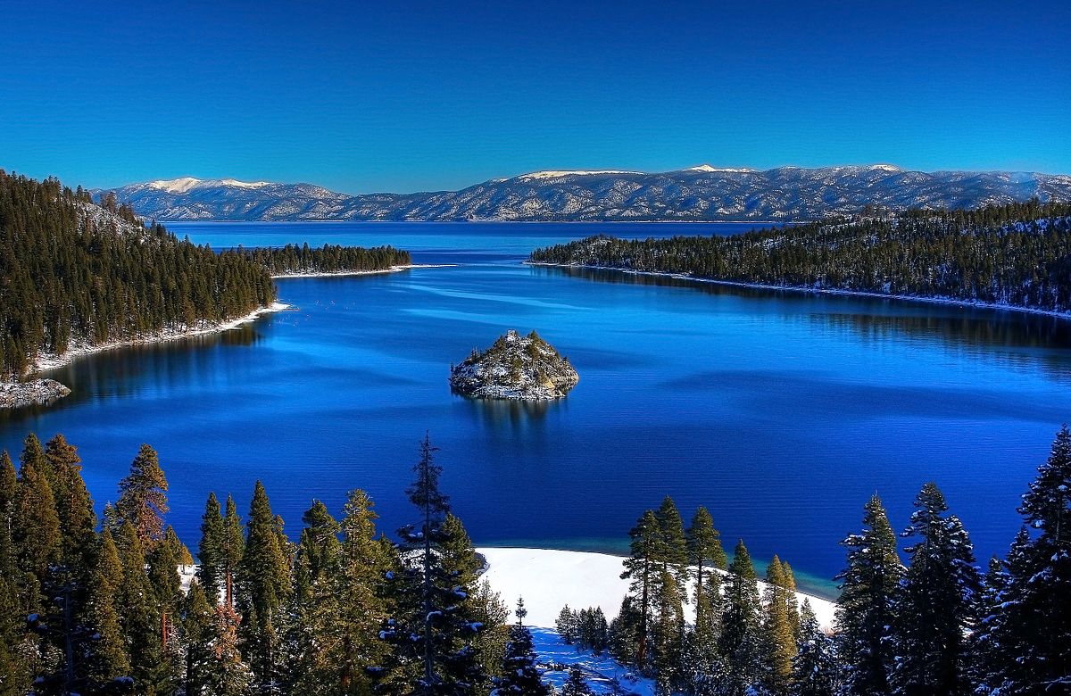 A stunning view of Lake Tahoe is presented, showcasing a deep blue lake surrounded by snow-capped mountains and a dense forest of evergreen trees. An island is visible in the center of the lake, highlighting the area's natural beauty, perfect for outdoor enthusiasts.